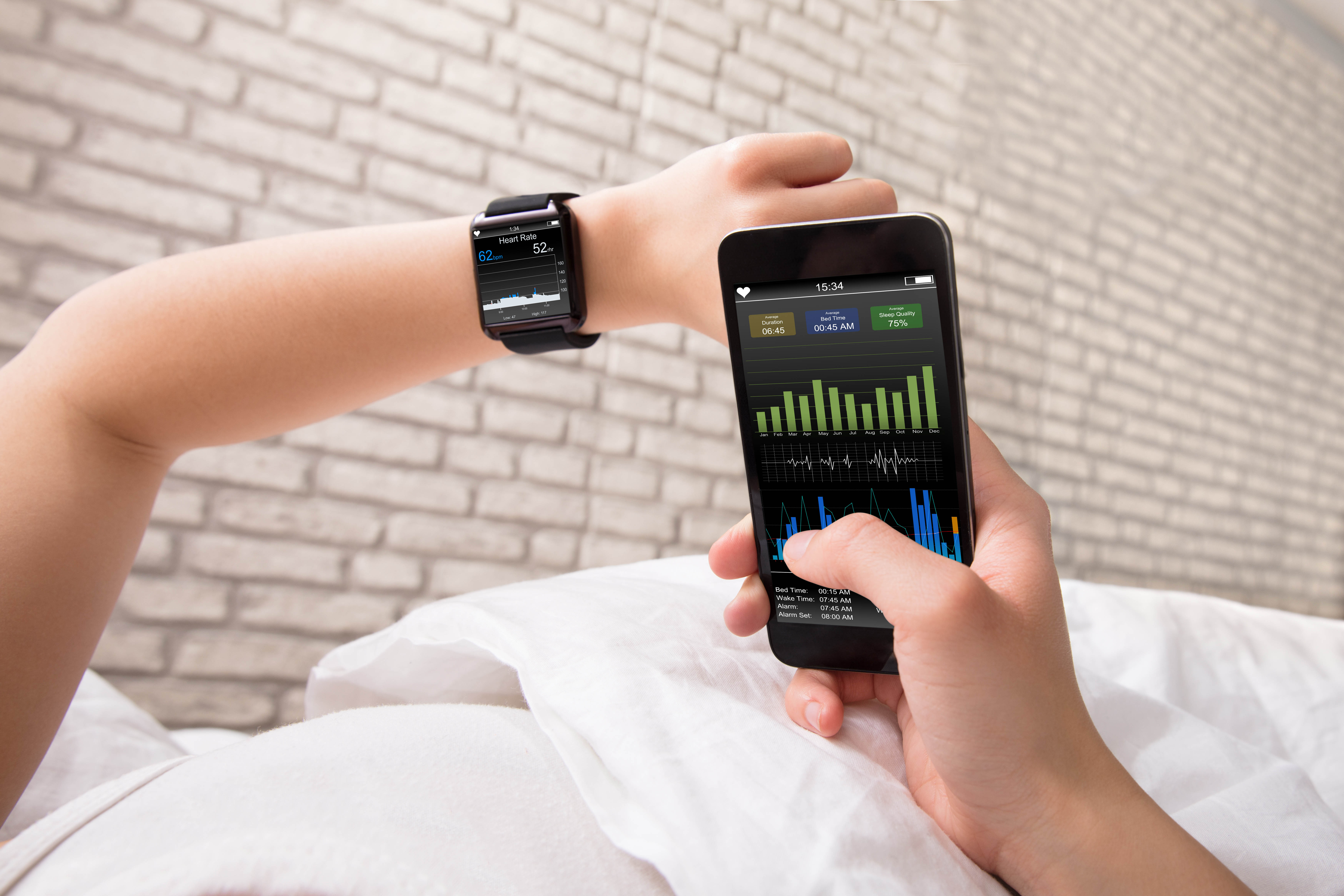 Close-up of woman's arm as she lays in bed. She's holding her one wrist with her smart watch up and is using her phone with the other hand to check her sleep stats.